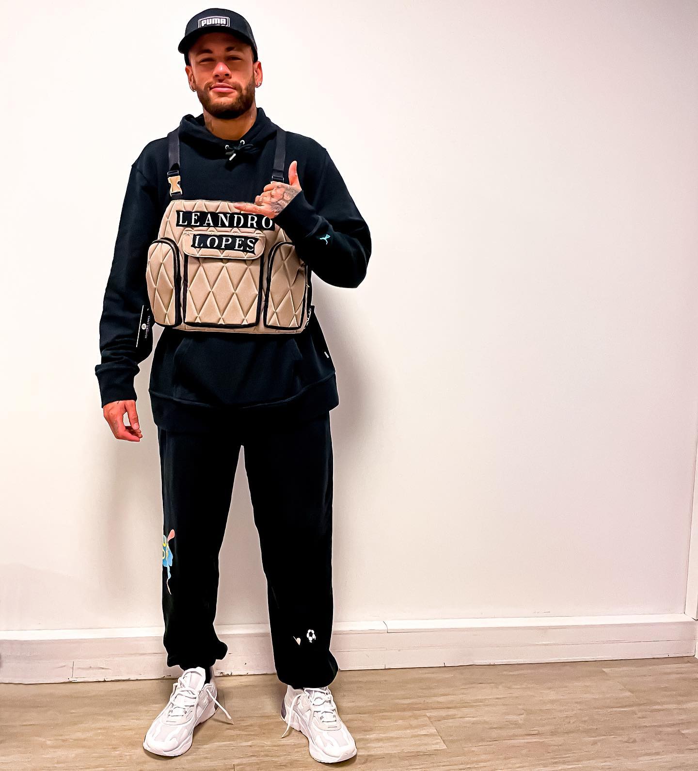 Person holding a beige backpack with 'Leandro Lopes' branding against a white wall.